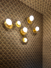 A fully wallpapered powder room turns a small space into an immersive jewel-box interior, with a dense brown, black, and cream pattern continuing across both the walls and ceiling. The illuminated All-Brass Virgo wall sconce by Sazerac Stitches features six globe bulbs, concave brass disc shades, and branching arms arranged like the Virgo zodiac constellation. This customizable sculptural lighting brings warm metallic contrast to maximalist bathroom decor and dramatic patterned walls. Discover a celestial l