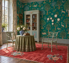 A richly layered sitting room combines teal Chinoiserie-style wallpaper, an aged floral rug, wide-plank wood floors, pale green metal chairs, and a skirted table topped with books and blue-and-white porcelain. The customizable Cream and Brass Virgo sconce by Sazerac Stitches traces its namesake zodiac constellation with six globe bulbs and sculptural branching arms. An inspired approach to statement wall lighting for grandmillennial, maximalist, traditional, and eclectic interiors. Explore celestial lightin
