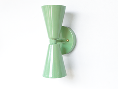 A soft sage-green double-cone wall sconce for a hallway, bathroom, bedroom, or mid-century modern room that wants gentle color and crisp lines. Shown straight on, the Dryades sconce pairs an upward and a downward sage cone at a narrow waist on a round sage backplate, with warm brass screw accents. Calm, considered, and easy to live with, its bowtie silhouette suits organic modern, colorful traditional, coastal, and English cottage spaces.
