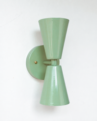A statement wall sconce for rooms that lead with soft, muted color and sculptural shapes. This angle of the Dryades sconce shows off its sage-green double-cone silhouette — one cone throwing light up, one down — meeting at a narrow waist on a round sage backplate with a brass screw accent. Calm, gentle, and a little retro, it's a soothing pick for hallways, baths, bedrooms, and mid-century or organic modern interiors that want color without commitment.