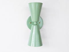 A soft, sculptural double-cone wall sconce for a hallway, bathroom, bedroom, or mid-century modern room that wants gentle color and crisp, two-directional light. The Dryades sconce pairs an upward and a downward sage-green cone at a narrow waist, mounted on a round sage backplate with a brass screw accent. Seen from an angle, its bowtie silhouette and wall projection read calm and considered — an easy, soothing pick for organic modern, colorful traditional, and English cottage spaces.