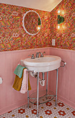 Close view of a colorful retro powder room with pink beadboard wainscoting, bold orange and green floral wallpaper, a round white mirror, and a vintage-style console sink with chrome legs. The Vista Green Thalia Sconce glows on the wall beside the mirror, adding a playful green accent to the pink and orange bathroom palette. This works for maximalist bathroom decor, 1970s-inspired wallpaper, colorful powder rooms, eclectic kids bathrooms, dopamine decor, and statement wall lighting.