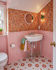 Wide view of a playful pink bathroom with beadboard walls, retro floral wallpaper, chrome console sink, round mirror, patterned hex tile floor, and layered towels in green, mustard, and blush pink. The Vista Green Thalia Sconce is lit on the wallpapered wall, bringing warm light and a sculptural pop of color to the space. This image fits colorful bathroom inspiration, grandmillennial powder room ideas, vintage-style sink styling, maximalist wallpaper, quirky wall sconces, and eclectic family bathroom decor.