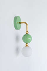 Calm, colorful vanity lighting that gives a bathroom or powder room a gentle point of view. Seen in profile, the Ida sconce shows off its curved brass arm reaching from a glossy sage-green backplate down to a stacked green sphere and frosted glass globe. The side view highlights the brass ball detailing and how far the fixture projects off the wall. A soft, soothing choice for colorful traditional, grandmillennial, English cottage, and organic modern interiors.