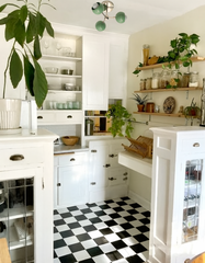 Vintage-inspired kitchen featuring the Loa Bicycle Club flushmount ceiling light in vista green and chrome, set against white cabinetry with glass-front doors, open wood shelving, and a classic black-and-white checkerboard floor. The bright space is accented with potted plants, jars, and natural wood details, blending retro charm with colorful modern lighting.
