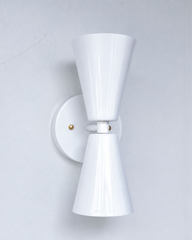 A statement wall sconce for rooms that lead with clean, sculptural shapes. This angle of the Dryades sconce shows off its glossy white double-cone silhouette — one cone up, one down — meeting at a narrow waist on a round white backplate, warmed by a brass screw accent. Crisp, versatile, and quietly confident, it's a soft classic for hallways, bathrooms, bedrooms, and mid-century or coastal interiors that want brass against a clean neutral.
