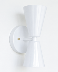 A clean, sculptural double-cone wall sconce for a hallway, bathroom, bedroom, or mid-century modern room that wants shape without loud color. The Dryades sconce pairs an upward and a downward glossy white cone at a narrow waist, mounted on a round white backplate with warm brass screw accents. Crisp and versatile, its bowtie silhouette reads well in mid-century, organic modern, and transitional spaces, with brass carrying the warmth.