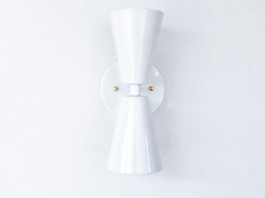 A clean, sculptural wall sconce for rooms that want shape and warmth without loud color. Seen from an angle, the Dryades sconce shows off its glossy white double-cone silhouette — one cone throwing light up, one down — meeting at a narrow waist on a round white backplate, with a brass screw accent. The three-quarter view highlights how it projects off the wall and its crisp mid-century lines. A versatile pick for hallways, baths, bedrooms, and organic modern or transitional interiors.