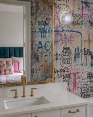This bathroom wraps its walls in a dense, multicolor graffiti mural wallpaper, full of scrawled tags, dates, and initials in blue, pink, and black. The Sid Sconce mounted beside the mirror keeps things polished with its white dome and gold spiked hardware, echoing the brass faucet and cabinet pulls elsewhere in the room. Through the doorway, a glimpse of a navy velvet headboard and pink pillows ties the bold wallpaper choice back to the rest of the house.
