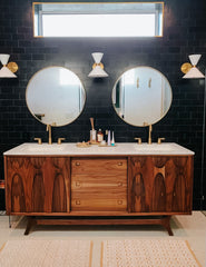 Modern white Clancy wall sconces with warm brass hardware mounted above a double bathroom vanity. The rich walnut cabinet and dark subway tile backsplash set the stage for these midcentury modern lights, perfect for layered bathroom lighting, contemporary home updates, or stylish vanity makeovers with timeless details.