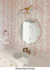White double cone wall sconce with brass arms and backplate mounted above a bathroom vanity, styled with a scalloped mirror and pink patterned wallpaper, a modern mid century inspired bathroom sconce for powder rooms, kids bathrooms, feminine interiors, and bright maximalist design with vintage brass details.