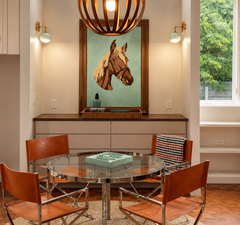 Symmetrical mint green and brass Loa wall sconces flank a bold equestrian horse art print, creating a statement lighting moment in an eclectic mid-century modern dining nook. Caramel leather sling chairs surround a round glass table on warm parquet wood floors, with built-in cabinetry and an oversized sculptural wood pendant adding vintage-inspired texture. A clean neutral backdrop keeps the focus on the colorful globe sconces, perfect for dining room lighting ideas, breakfast nook wall lighting, and modern