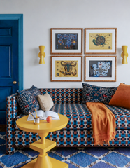 This living room commits to pattern mixing without apology: a geometric woven sofa, a diamond-print rug, floral and velvet pillows, and a four-panel gallery wall of surreal artwork all sharing one wall. A pair of yellow Dryades Sconces flank the art symmetrically, their pinched hourglass shape adding a graphic note that echoes the yellow pedestal side table below. A deep blue door in the corner ties the whole palette together, proving a room can clash on purpose and still feel intentional.
