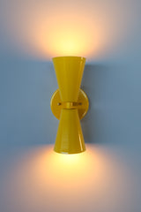 The Dryades Sconce in a vibrant, high-gloss yellow finish shown fully illuminated against a cool-toned wall, casting a warm glow both upward and downward. This bold, double-cone wall light features a clean mid-century modern silhouette with a matching yellow backplate and subtle brass hardware details. The cheerful color and directional light pattern make it perfect for hallways, bathrooms, or accent walls in maximalist, eclectic, or modern interiors. A playful yet functional wall fixture ideal for adding p
