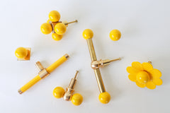 A lineup of yellow cabinet hardware in six different shapes, from a multi-ball cluster knob and a simple single ball knob to a bar pull, a long double-ball pull, a small gumball knob, and a scalloped flower knob, all finished with brass posts and backplates. A good one-stop reference for mixing pull styles across different drawer and cabinet sizes without losing the color story.