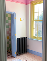Bright hallway with pale walls, pink crown detail, powder blue door and baseboards, and sunshine yellow window trim; Sazerac Stitches Abeille wall light in yellow with white globe mounted beside a black cast-iron radiator; natural light on wide plank wood floors, glimpse into an adjacent bath with pastel checkerboard tiles and vintage mosaic floor; playful color-blocked interior, compact low-profile fixture ideal for narrow walls and small spaces; handmade customizable lighting from New Orleans.