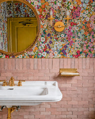 A playful, high end powder room moment with blush pink zellige tile wainscoting and a dense, colorful floral wallpaper that feels like modern grandmillennial decor. A round brass mirror and brass sink fittings add warmth, while the Sazerac Stitches Adele two light wall sconce in sunny yellow and brass brings the perfect pop of color. A great idea for statement bathroom lighting, floral wallpaper bathrooms, pink tile bathrooms, boutique hotel powder rooms, eclectic interiors, and maximalist decor. Handmade i