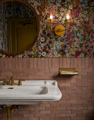 Same space, different mood. With the lights on, the yellow and brass Adele two light sconce casts a warm, candle like glow over the pink zellige tile and vibrant floral wallpaper, making the whole bathroom feel cozy and glamorous. This is a great reference image for colorful powder rooms, brass bathroom lighting, statement sconces, maximalist wallpaper, and cheerful guest bath design. Handmade in New Orleans.
