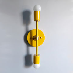 right yellow and brass Medium Thalia Sconce with double globe bulbs, bold color-pop mid century modern wall sconce in a vibrant color-drenched palette, modern vertical wall light with round backplate, perfect for maximalist interiors, kids room lighting, playful nursery decor, sunny hallway wall light, or statement bathroom vanity lighting by Sazerac Stitches