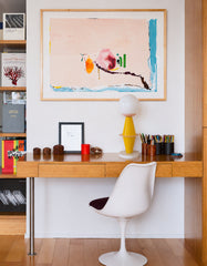 An adult creative workspace styled with the yellow and blush pink Totem Lamp on a warm wood mid century modern desk. The room mixes clean modernist furniture with colorful art, including a large abstract painting, built-in bookshelves, pencil cups, small collected objects, and a white tulip-style desk chair. The Totem Lamp adds playful sculptural lighting without feeling childish, thanks to its glossy yellow stacked base, soft blush pink details, white globe shade, and marble foot. This image fits an adult 