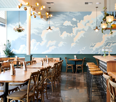 An airy restaurant dining room with a hand-painted cloud mural in soft pastel blues, dark green wainscoting below, and a Large Octavia Chandelier in brass glowing warm overhead near the sloped ceiling. Rich wood farmhouse tables, vintage bentwood chairs, and polished concrete floors keep the space feeling natural and unfussy, cozy and atmospheric without trying too hard. A breeze-block bar and trailing greenery round out the room in the background.
