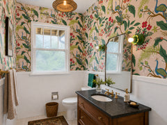 Wide view of a colorful bathroom with peacock and botanical wallpaper, white tile wainscoting, a warm wood vanity, dark stone countertop, brass faucet, black framed mirror, and Aged Green Octavia Sconces used as statement bathroom lighting. The leafy floral shape of the sconce blends into the tropical wallpaper while the glowing globe bulb adds a soft, playful accent. This works for maximalist bathrooms, grandmillennial powder rooms, tropical wallpaper, peacock decor, colorful vanity lighting, and eclectic 