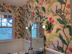 Close view of the Aged Green Octavia Sconce mounted on bold peacock wallpaper in a colorful bathroom. The green flower-like fixture and glowing white globe bulb sit against a playful botanical wallcovering with leaves, branches, and blue peacocks, creating a layered tropical look. The room also includes a black framed mirror, white tile wainscoting, wood vanity, and brass accents. A strong fit for searches around floral wall sconces, green bathroom lighting, tropical powder rooms, maximalist wallpaper, and 