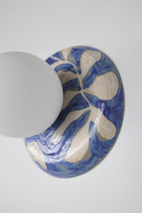 Closeup detailed image of Handmade one-of-one watercolor blue and white leafy ceramic globe sconce with a matte white round globe shade mounted on a white wall, glossy botanical brushstroke pattern, sculptural artisan wall light for bedside lighting, hallway accent lighting, entryway statement, and bathroom vanity style spaces in modern, coastal, and grandmillennial decor