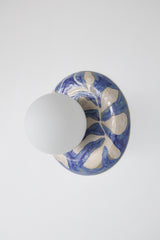 Side view of a one-of-one handmade ceramic globe wall sconce showing the sculptural curved ceramic backplate with glossy blue and white leafy watercolor pattern and a matte white globe shade, artisan pottery lighting with a clean modern profile for designer interiors, bedside sconces, hallway lighting, and statement wall fixtures
