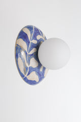 Handmade one-of-one watercolor blue and white leafy ceramic globe sconce with a matte white round globe shade mounted on a white wall, glossy botanical brushstroke pattern, sculptural artisan wall light for bedside lighting, hallway accent lighting, entryway statement, and bathroom vanity style spaces in modern, coastal, and grandmillennial decor