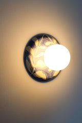 Close wall view of a lit one-of-one handmade ceramic globe sconce with a matte white globe and a glossy blue and white leafy watercolor botanical backplate, warm soft ambient light, artisan pottery wall fixture for elevated bedroom lighting, entryway accents, hallway glow, and designer statement lighting