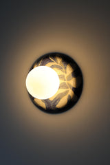 Lit handmade one-of-one ceramic wall sconce with a matte white globe shade and a glossy blue and white watercolor leafy pattern, warm ambient glow washing the wall, sculptural artisan lighting for modern, coastal, and eclectic interiors, ideal for bedside wall lamps, hallway lighting, and statement accent fixtures