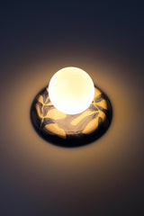 Close wall view of a lit one-of-one handmade ceramic globe sconce with a matte white globe and a glossy blue and white leafy watercolor botanical backplate, warm soft ambient light, artisan pottery wall fixture for elevated bedroom lighting, entryway accents, hallway glow, and designer statement lighting