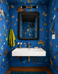 A dramatic jewel-box powder room wrapped in saturated cobalt blue walls covered with colorful butterflies and birds, creating an immersive maximalist wallpaper moment. A sculptural black statement mirror with a chunky ribbed frame sits above a crisp white wall-mounted sink, finished with a warm brass faucet and matching brass plumbing. Centered over the mirror is the Sazerac Stitches Janus sconce in black and brass, a mid-century modern double-cone wall light that adds sharp graphic contrast against the viv
