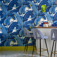 A chartreuse shell sconce popping against a blue banana-leaf wallpaper — the exact kind of high-voltage color clash that makes a home bar unforgettable. The Coquille from Sazerac Stitches is a glossy scallop-shell wall light with a warm brass fitting, here in electric lime-green against a classic tropical palm-leaf print. The room layers it up: a curved white bar, blue-and-white leopard-print bar stools with brass legs, chartreuse baseboards, cobalt glassware, and terrazzo floors. Full-tilt maximalism with 