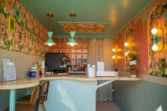 Color-drenched micro café interior with three mint green Clancy pendants over a curved stone counter, paired with Thalia wall sconces glowing against pink floral wallpaper; aqua tile wainscoting, brass accents, glass-block partition, and a scalloped ceiling inset create a playful midcentury look; Sazerac Stitches powder-coated steel shades with raw brass hardware, handmade in New Orleans; design credit @brick_and_barley for @brighterdayscoffee.