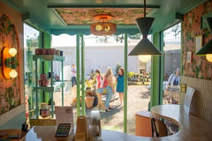 Bright café looking toward mint green framed glass doors with sunlight and shoppers outside; mint Clancy pendants with globe bulbs hang above the counter, Thalia double-globe sconces warm the patterned pink walls, open shelving with mugs, rattan ceiling fixture, and curved countertop; bold pastel palette, midcentury modern vibe; Sazerac Stitches customizable powder-coated lighting handmade in New Orleans; design by @brick_and_barley at @brighterdayscoffee.