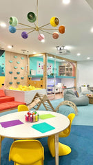 Colorful kids’ playroom with rainbow Arcenciel chandelier by Sazerac Stitches, yellow chairs, art supplies, climbing wall, and creative play zones.