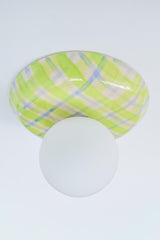 Angled studio view of a glossy plaid ceramic wall sconce in lime green, cream, and pale blue with a matte white globe shade, emphasizing the curved ceramic silhouette and reflective glaze, a colorful statement globe sconce for dopamine decor, eclectic modern interiors, playful wall lighting, and design-forward accent lighting in entryways, bedrooms, and powder rooms.