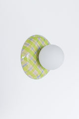 Angled studio view of a glossy plaid ceramic wall sconce in lime green, cream, and pale blue with a matte white globe shade, emphasizing the curved ceramic silhouette and reflective glaze, a colorful statement globe sconce for dopamine decor, eclectic modern interiors, playful wall lighting, and design-forward accent lighting in entryways, bedrooms, and powder rooms.