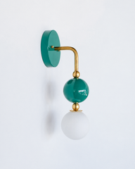 A statement wall sconce for rooms that lead with rich, saturated color. This angle of the Ida sconce puts the bold round teal-green backplate front and center, with a brass arm curving down to a glossy green sphere and frosted glass globe. Deep, jewel-toned, and quietly confident, it's a sophisticated pick for colorful bathrooms, powder rooms, bedrooms, and eclectic spaces that want one soulful, standout detail.