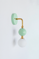 A soft mint wall sconce for a nursery, girl's bedroom, or pastel powder room that wants gentle, sweet color. Shown straight on, the Ida sconce stacks a glossy mint powder-coated sphere over a frosted glass globe, joined by warm brass hardware and a round mint backplate. The head-on view shows how the spheres line up into a tidy vertical drop. Pretty and calming, it's a standout for grandmillennial, coastal traditional, and playful kids' spaces.