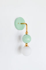 Sweet, calming vanity lighting that gives a bathroom, nursery, or girl's bedroom a gentle point of view. Seen in profile, the Ida sconce shows off its curved brass arm reaching from a glossy pastel mint backplate down to a stacked mint sphere and frosted glass globe. The side view highlights the brass ball detailing and how far the fixture projects off the wall. A soft, pretty choice for grandmillennial, English cottage, and colorful traditional interiors.