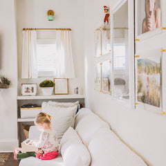 Cozy neutral living room featuring a mint green Loa wall sconce with brass backplate mounted above a bright window and white curtains on a brass rod. Soft modern styling with a white sofa, framed photo wall, ledge shelf with family photos, and warm natural light. Great for searches like modern wall lighting, mint wall sconce, brass sconce, Scandinavian living room decor, and airy neutral interiors.