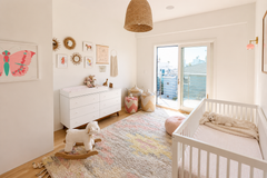 Bright neutral nursery with soft white walls, a white crib, and a cozy textured rug in pastel tones. A blush pink Loa wall sconce with brass hardware adds a warm pop of color near the window and floor length curtains. Mid-century modern white dresser with wood legs, woven baskets, playful gallery wall art, and a rattan pendant light create an airy Scandinavian boho nursery vibe that feels modern, calm, and design-forward.