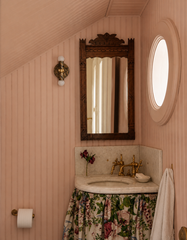 Pale pink beadboard powder room with a marbled Thalia wall sconce in olive and cream tones with brass hardware, styled beside an antique carved wood mirror. Curved corner marble sink with a floral fabric skirt, warm unlacquered brass faucet, and soft vintage cottage details under a slanted ceiling. Grand millennial bathroom inspiration with coastal and English country vibes, eclectic traditional style, statement bathroom lighting, decorative wall sconce for a small powder room.