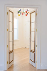 Rainbow Prism Chandelier with twelve colorful powder-coated steel globes on a brass frame, installed in a bright minimalist entryway with white walls, tall double French doors, and light wood floors — a midcentury modern statement light fixture that adds bold color and playful design to transitional spaces, perfect for dining rooms, playrooms, or hallways in eclectic and contemporary homes.