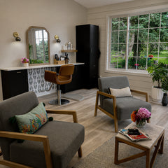 Bright, modern hair stylist studio with a pair of compact Loa wall sconces in brushed steel and brass, mounted on a warm beige wall above a salon styling station. The small dome-and-globe sconces add soft, focused task lighting around a gold-framed mirror, black cabinetry, and a clean countertop with hair products, balancing mixed metals with matte black finishes. Large picture window and white shiplap walls bring in natural light, while mid-century inspired lounge chairs and a neutral rug keep the space ai