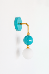 A statement wall sconce for rooms that lead with saturated blue-green color. This angle of the Ida sconce puts the bold round turquoise backplate front and center, with a brass arm curving down to a glossy teal sphere and frosted glass globe. Rich, watery, and confident, it's a spirited pick for coastal bathrooms, powder rooms, kids' spaces, and colorful traditional rooms that want one joyful, standout detail.