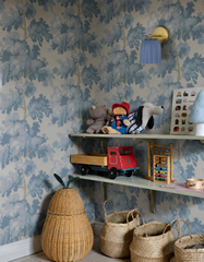 Dusty blue tree wallpaper sets a soft, storybook mood in this kids room, where open wood shelving holds vintage-style wooden toys, a stuffed animal lineup, and a stack of woven seagrass baskets below. The Marcella Sconce in unglazed blue picks up the wallpaper's exact tone, proving lighting can match a room's palette instead of standing apart from it. A calm, collected take on kids room wallpaper and playroom styling.